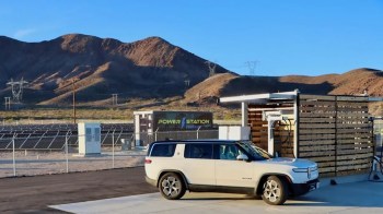 A white electric vehicle is parked near a charging station with solar panels and mountains in the background.