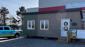 A small green and brown tiny home with a wreath on the door, displayed outside a building, alongside a blue pickup truck.