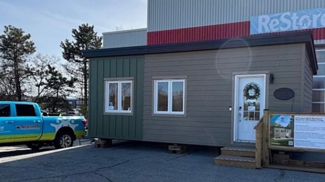 A small green and brown tiny home with a wreath on the door, displayed outside a building, alongside a blue pickup truck.