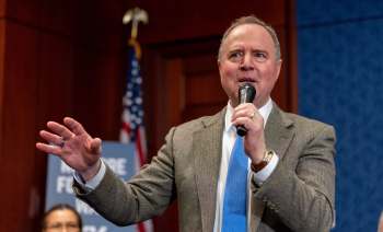 A man in a suit speaks into a microphone during a press conference, gesturing with his hand.