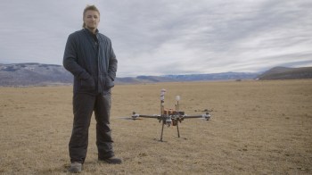 A person stands in an open field next to a drone, with mountains visible in the background under overcast skies.