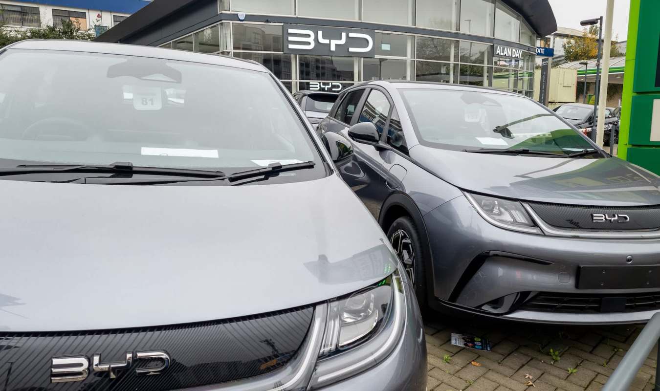 Two grey BYD electric cars are parked in front of a dealership with the BYD logo prominently displayed.