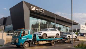 A transport truck unloading two electric vehicles in front of a modern BYD dealership.