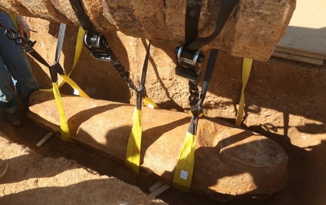 A large, ancient stone artifact being lifted from the ground using yellow straps and pulleys.