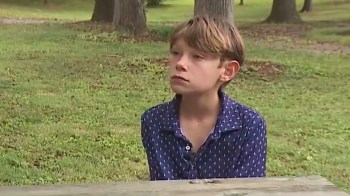 A young boy with short hair sits at a wooden table in a grassy park, looking serious and contemplative.