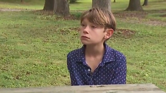 A young boy with short hair sits at a wooden table in a grassy park, looking serious and contemplative.
