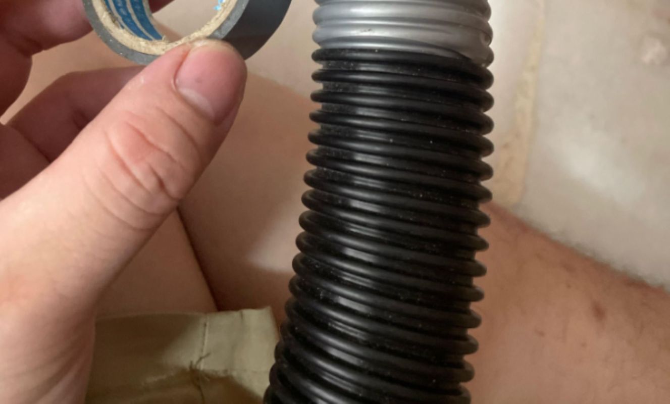 A hand holding a roll of tape next to a black, ribbed hose.