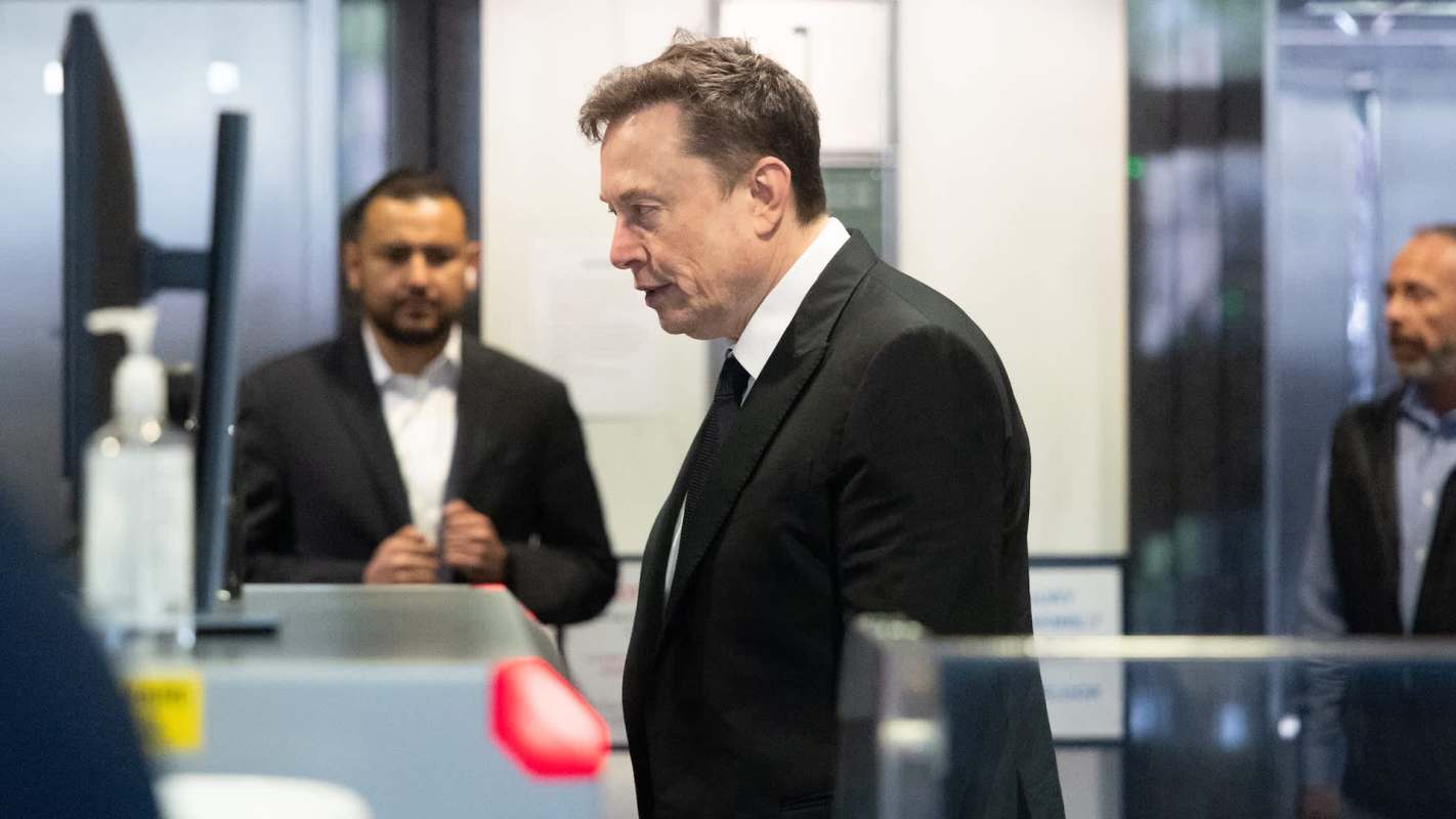 Elon Musk walks past a security station with two other men in the background.