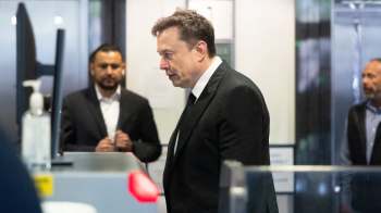Elon Musk walks past a security station with two other men in the background.