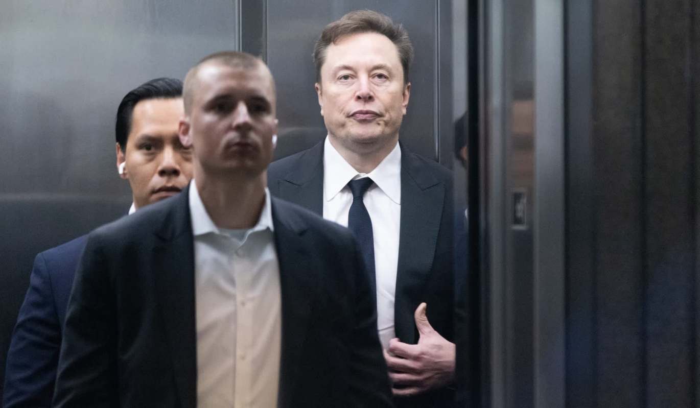 Elon Musk and two other men in suits stand inside an elevator, with Musk looking sternly at the camera.