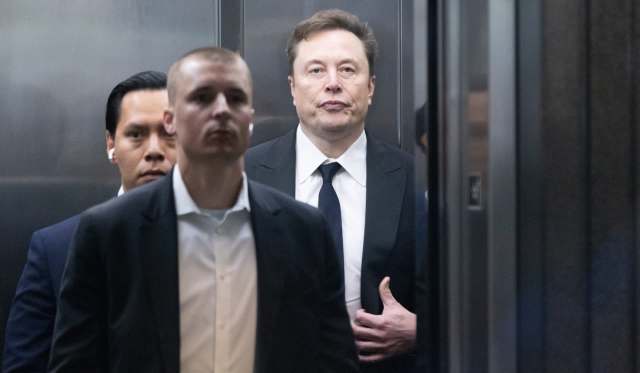 Elon Musk and two other men in suits stand inside an elevator, with Musk looking sternly at the camera.