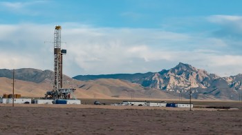 A drilling rig is set in a rugged landscape with mountains and clear skies in the background.