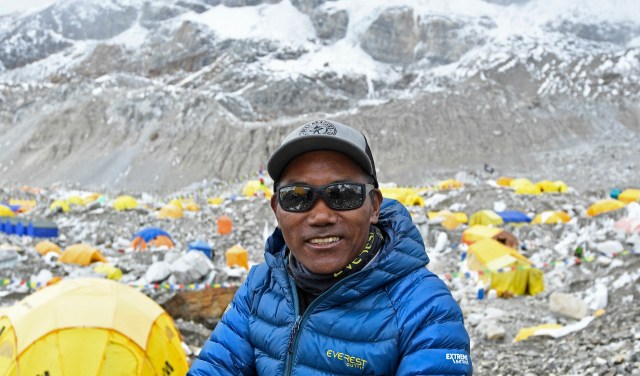A man in sunglasses smiles in front of a colorful campsite at the base of a snow-capped mountain.