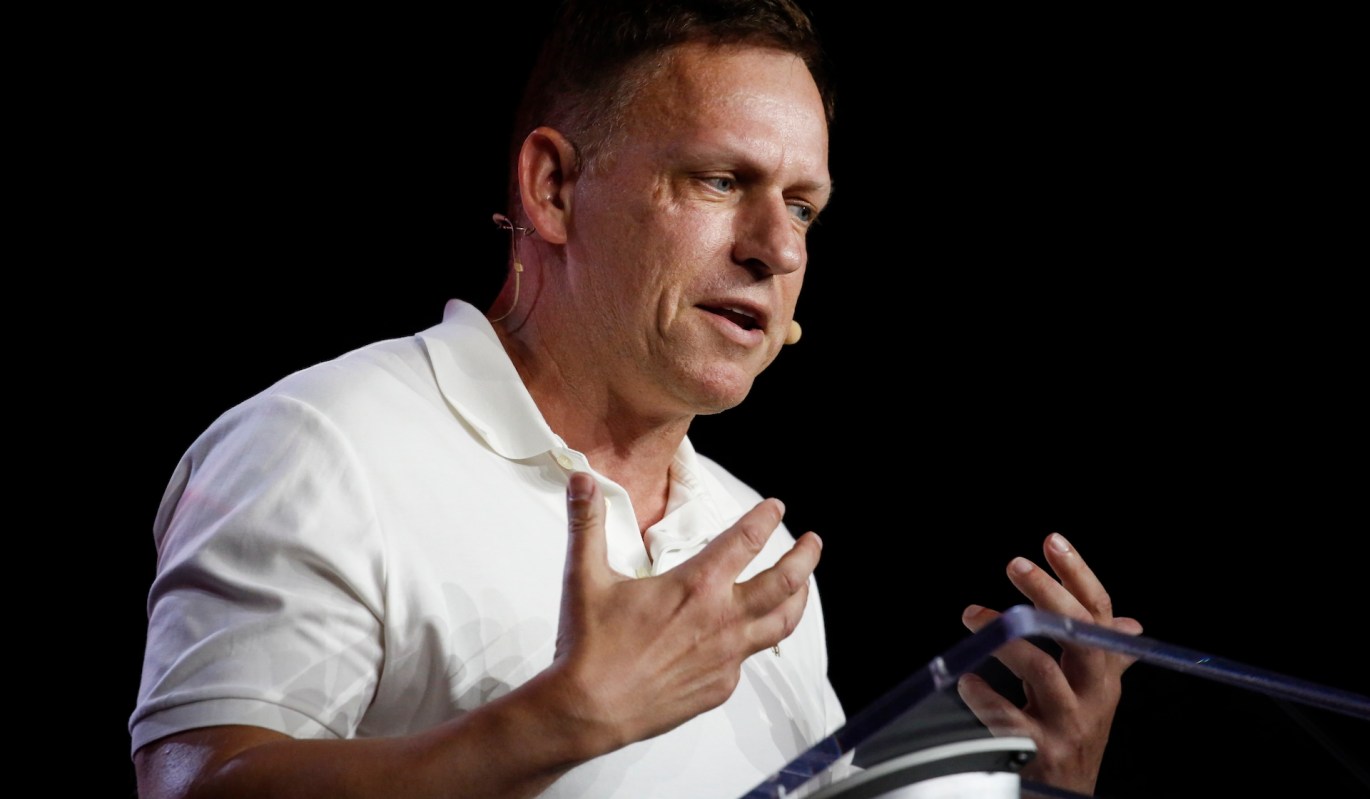 Peter Thiel, in a white polo shirt, speaks at a podium.