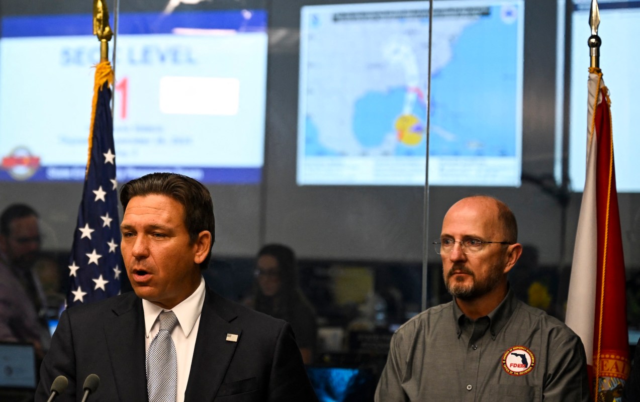 Florida Gov. Ron DeSantis speaks at a podium while another man listens, with a weather map displayed in the background.