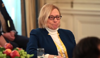 Maine Governor Janet Mills, wearing glasses, looks contemplative while seated at a table with fruit, wearing a blue jacket.