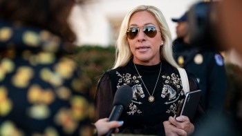 Marjorie Taylor Greene, with long blonde hair, wearing sunglasses and a black top, speaks into a microphone outdoors.