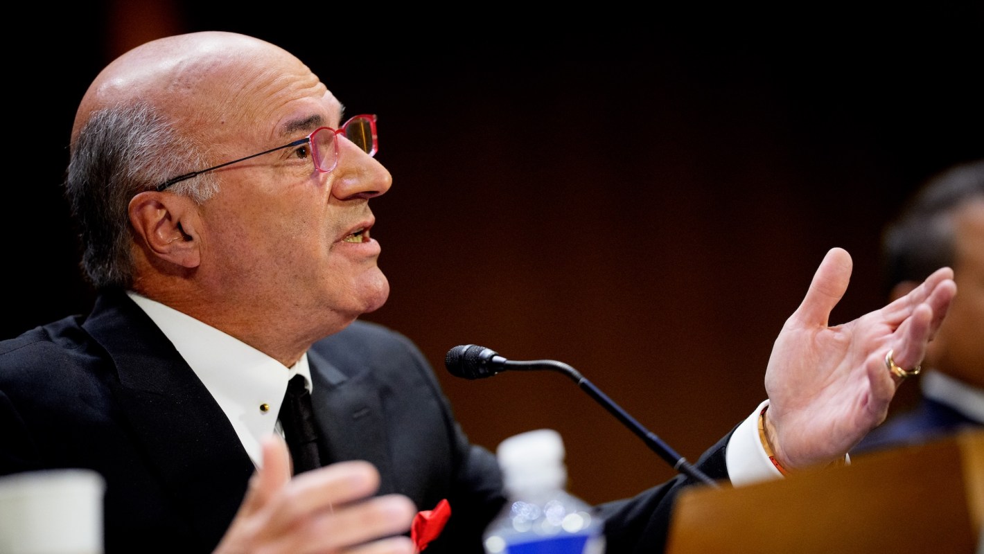 Kevin O'Leary in a suit gestures while speaking at a hearing, wearing pink glasses and a red pocket square.