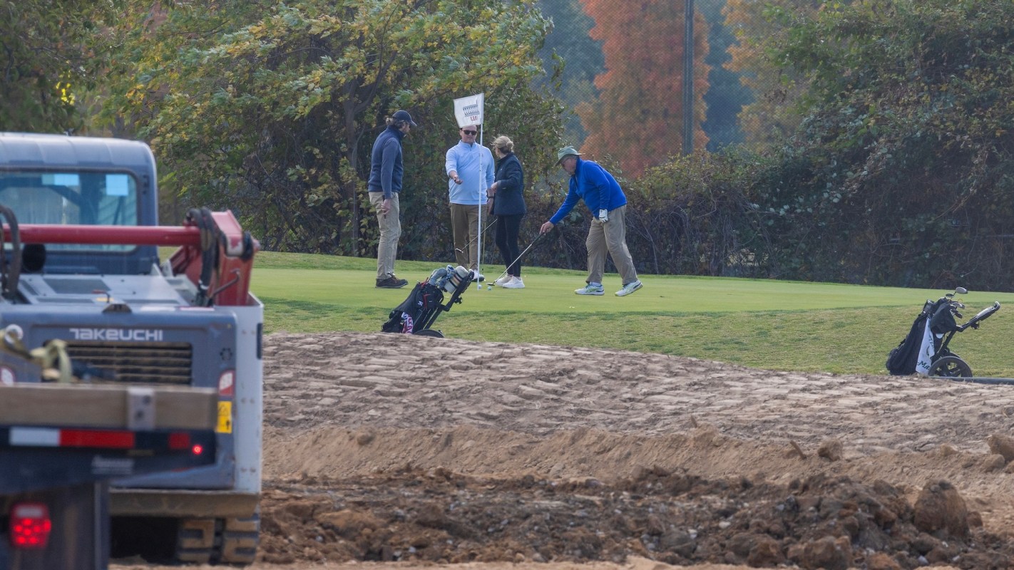 A group of golfers on a green, with construction equipment and golf bags in the foreground.
