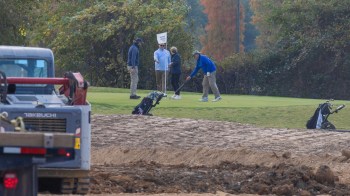 A group of golfers on a green, with construction equipment and golf bags in the foreground.