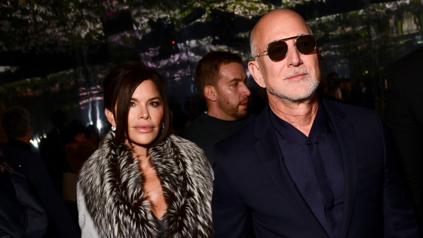Lauren Sánchez Bezos, in a fur coat, and Jeff Bezos, in sunglasses, attend an event in front of a dark background.