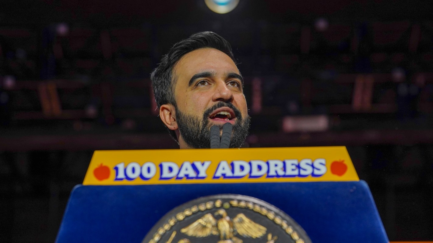 NYC Mayor Zohran Mamdani speaks passionately at a podium labeled "100 Day Address" with the city's seal in the foreground.