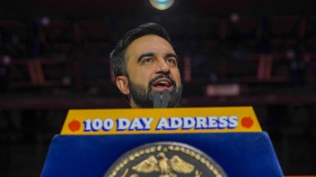 NYC Mayor Zohran Mamdani speaks passionately at a podium labeled "100 Day Address" with the city's seal in the foreground.