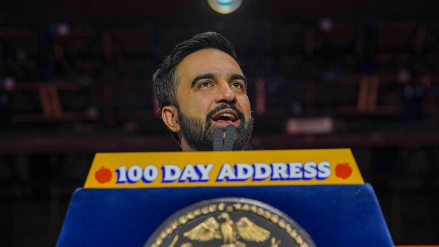 NYC Mayor Zohran Mamdani speaks passionately at a podium labeled "100 Day Address" with the city's seal in the foreground.