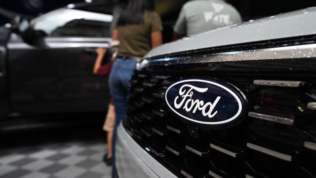 A close-up of a Ford emblem on a vehicle with raindrops, with people and a car in the background.