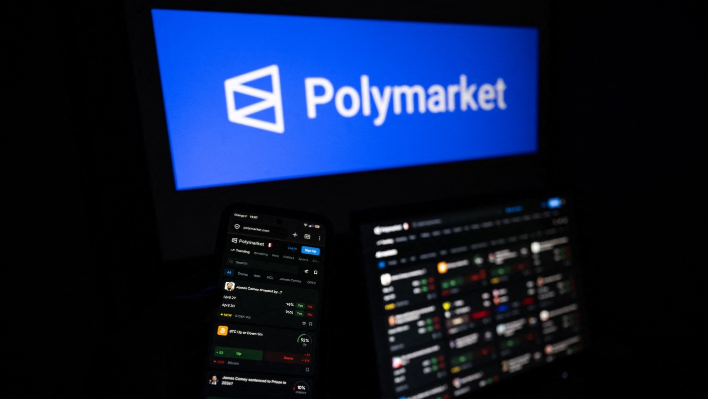 A smartphone displaying the Polymarket platform, with a logo in the background on a computer screen.