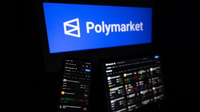 A smartphone displaying the Polymarket platform, with a logo in the background on a computer screen.