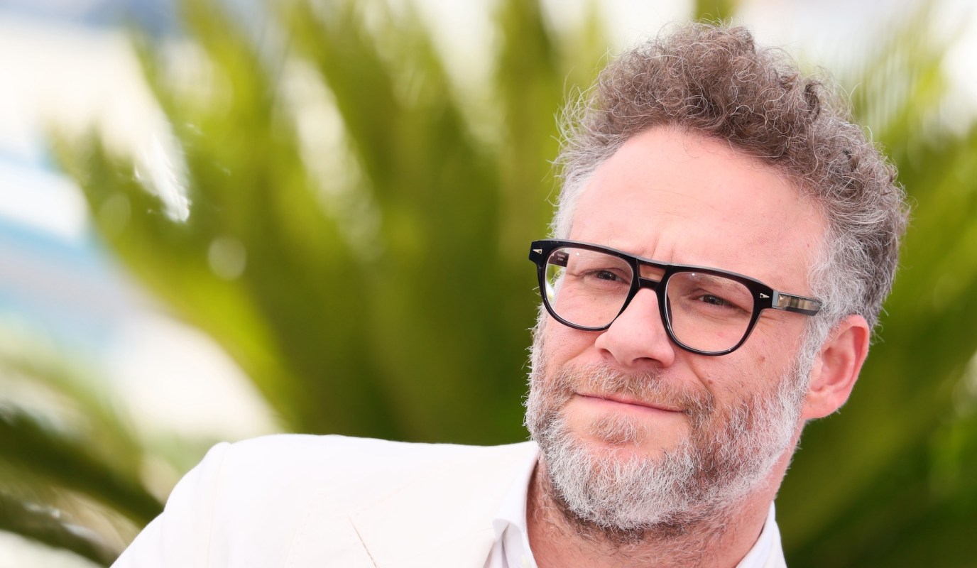 Seth Rogen, with curly hair and glasses, poses outdoors against a leafy backdrop.