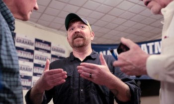 U.S. Senate candidate Graham Platner, with a beard, gestures while talking with two others in a room.