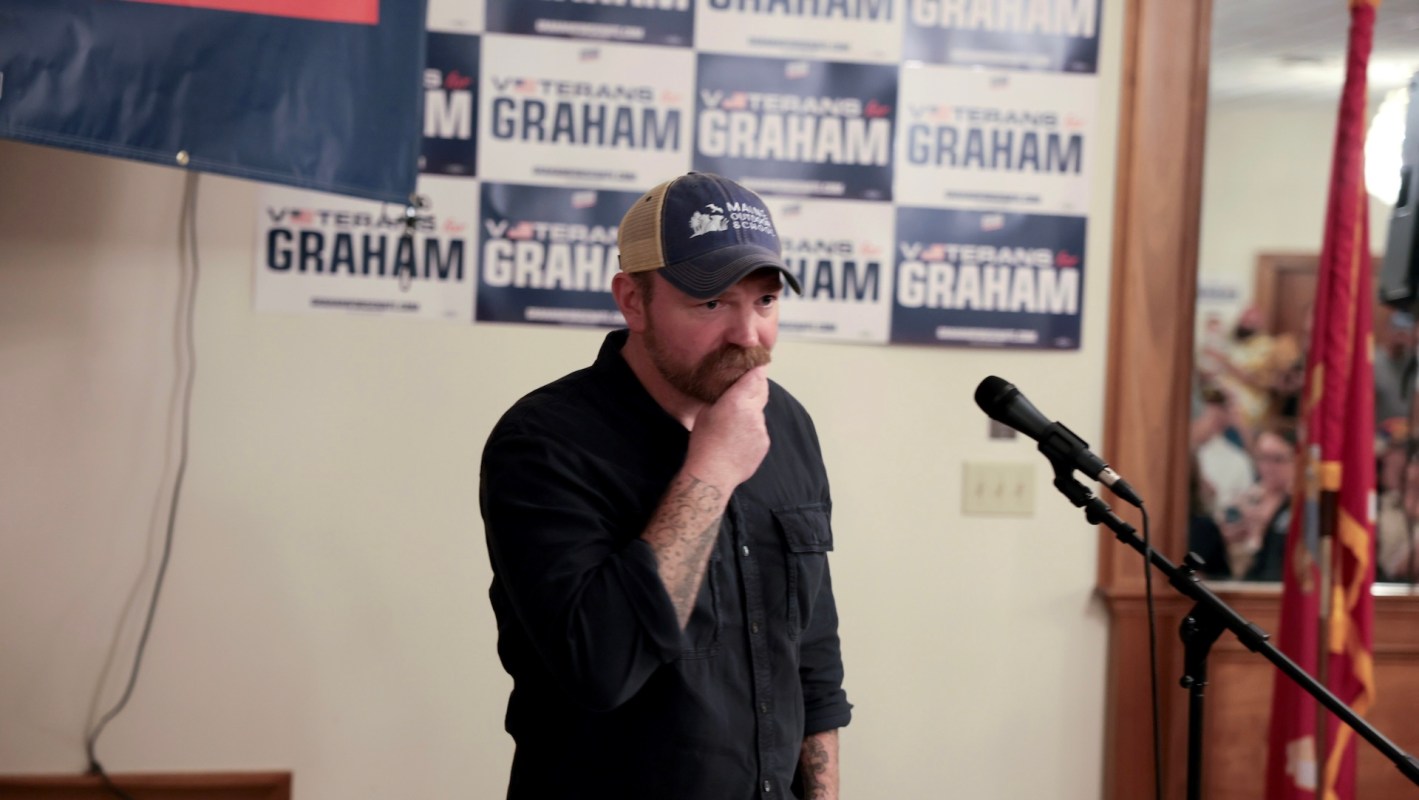 Graham Platner with a mustache stands near a microphone, looks down in front of campaign signs for his U.S. Senate campaign.