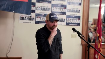 Graham Platner with a mustache stands near a microphone, looks down in front of campaign signs for his U.S. Senate campaign.