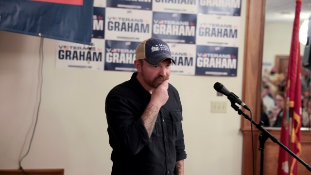 Graham Platner with a mustache stands near a microphone, looks down in front of campaign signs for his U.S. Senate campaign.
