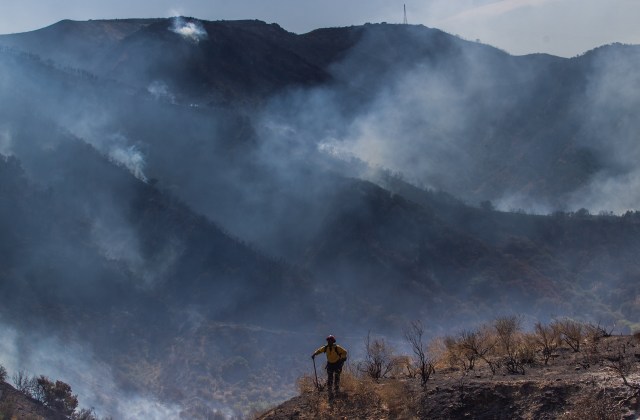 A firefighter surveys a smoky, burned landscape on a mountainous terrain.