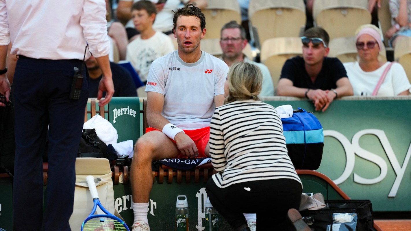 Norwegian tennis player Casper Ruud sits on a chair, receiving assistance from a woman while spectators watch.