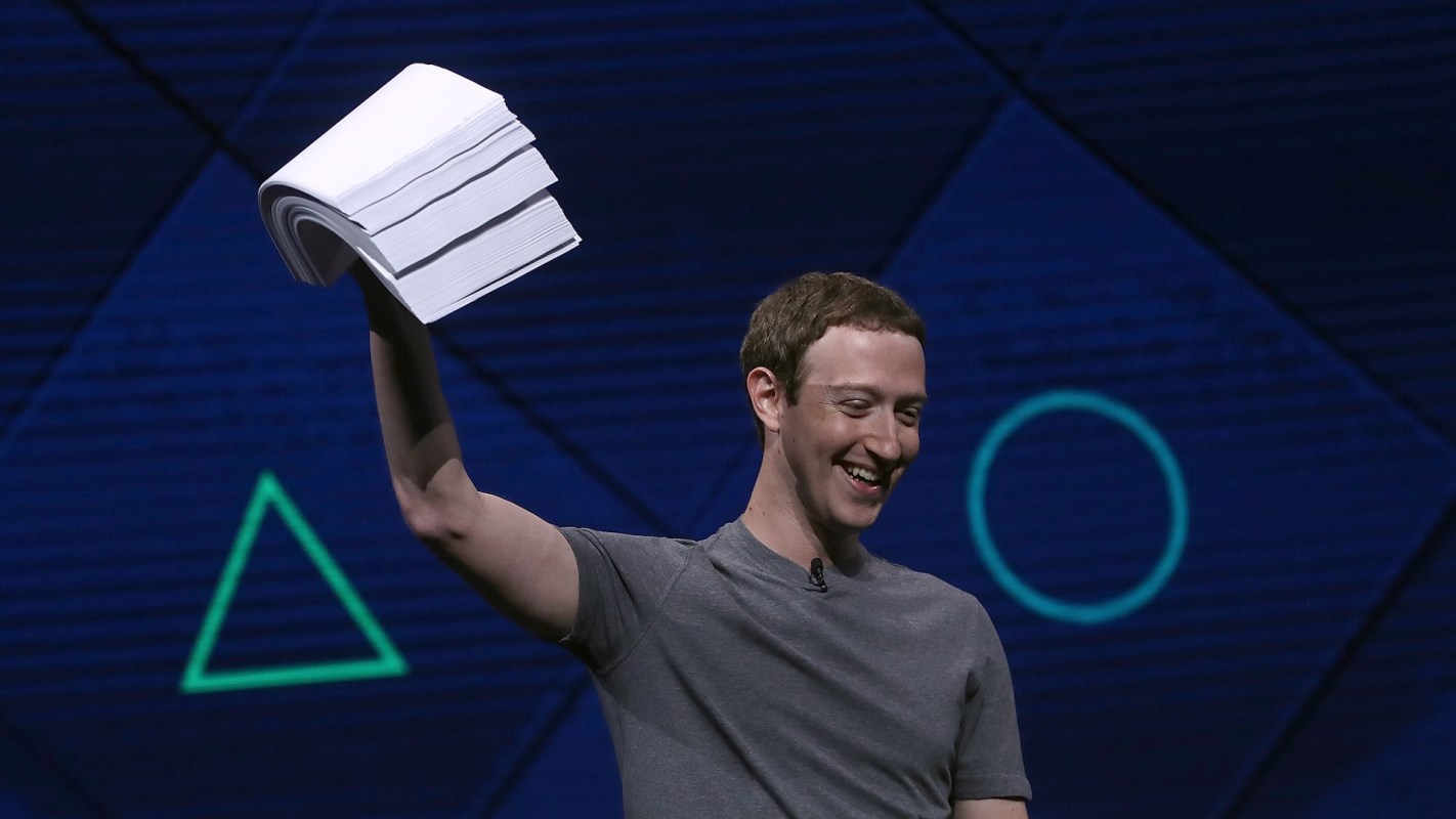 A smiling Mark Zuckerberg in a gray shirt holds up a stack of papers on a stage with geometric patterns in the background.