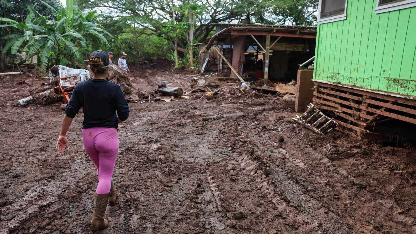 A person walks through muddy ground near a green structure, with debris visible in a damaged environment.