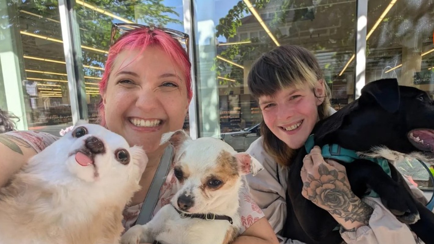 Two smiling people pose for a selfie with three small dogs outside a store on a sunny day.