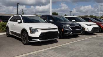 A lineup of various KIa SUVs parked side by side in a dealership lot under a partly cloudy sky.