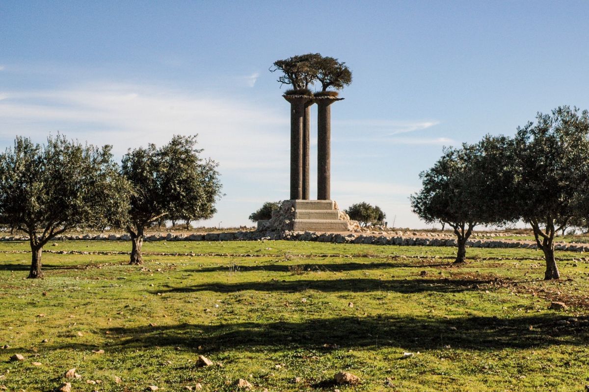 A sculpture featuring two columns topped with a tree, surrounded by olive trees in a grassy landscape.