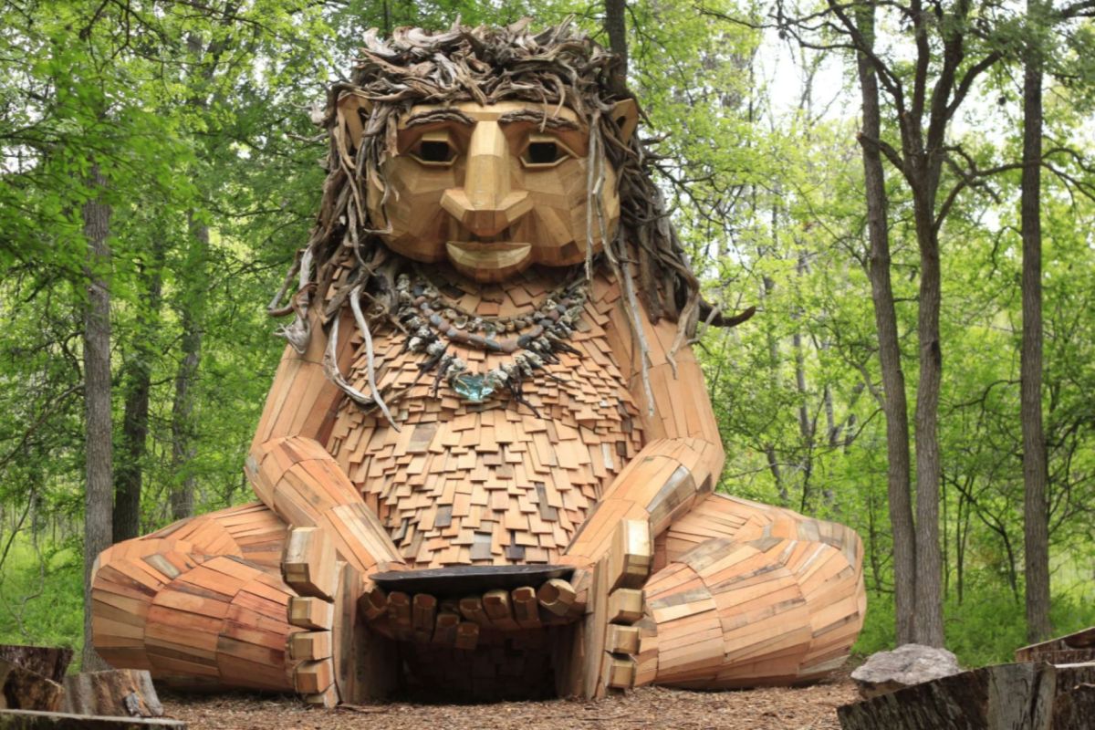 A large wooden sculpture of a seated figure with leafy hair, surrounded by trees in a forested area.