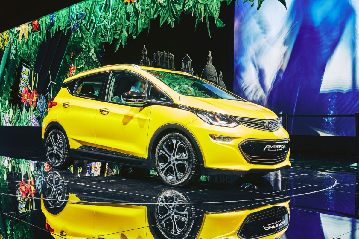 A bright yellow electric car displayed on a reflective surface with a floral backdrop.