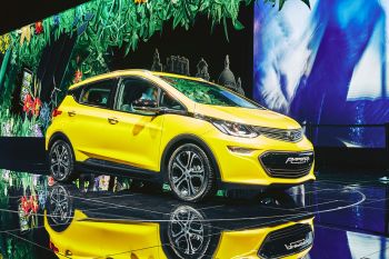 A bright yellow electric car displayed on a reflective surface with a floral backdrop.