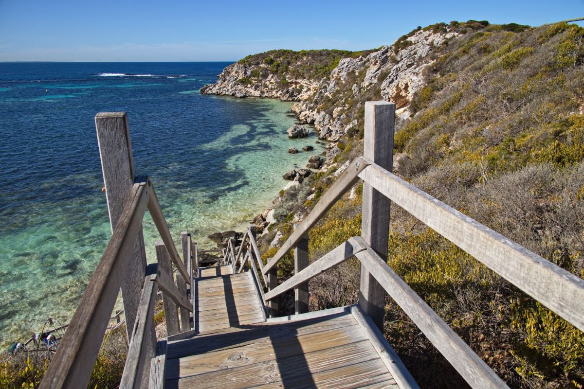 A wooden staircase leads down to a clear blue sea with rocky shores and green hills in the background.
