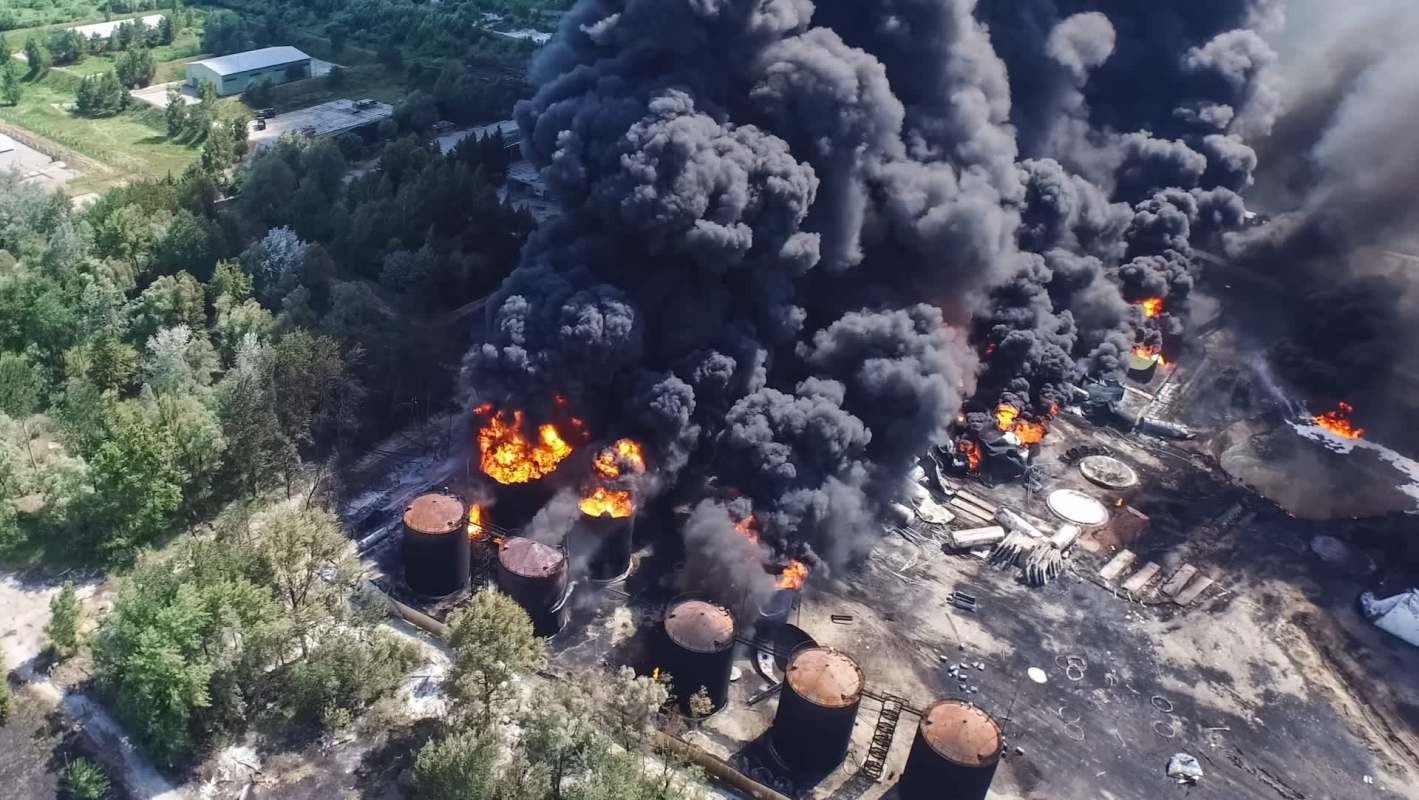 An aerial view of a large industrial fire with thick black smoke and flames rising from storage tanks.
