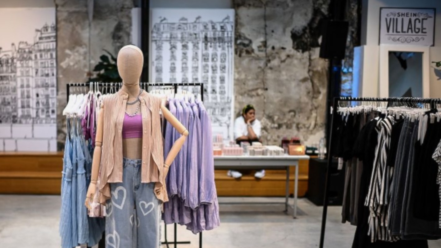 A mannequin displays casual apparel in a Shein retail store, with hanging clothes and a seated employee in the background.
