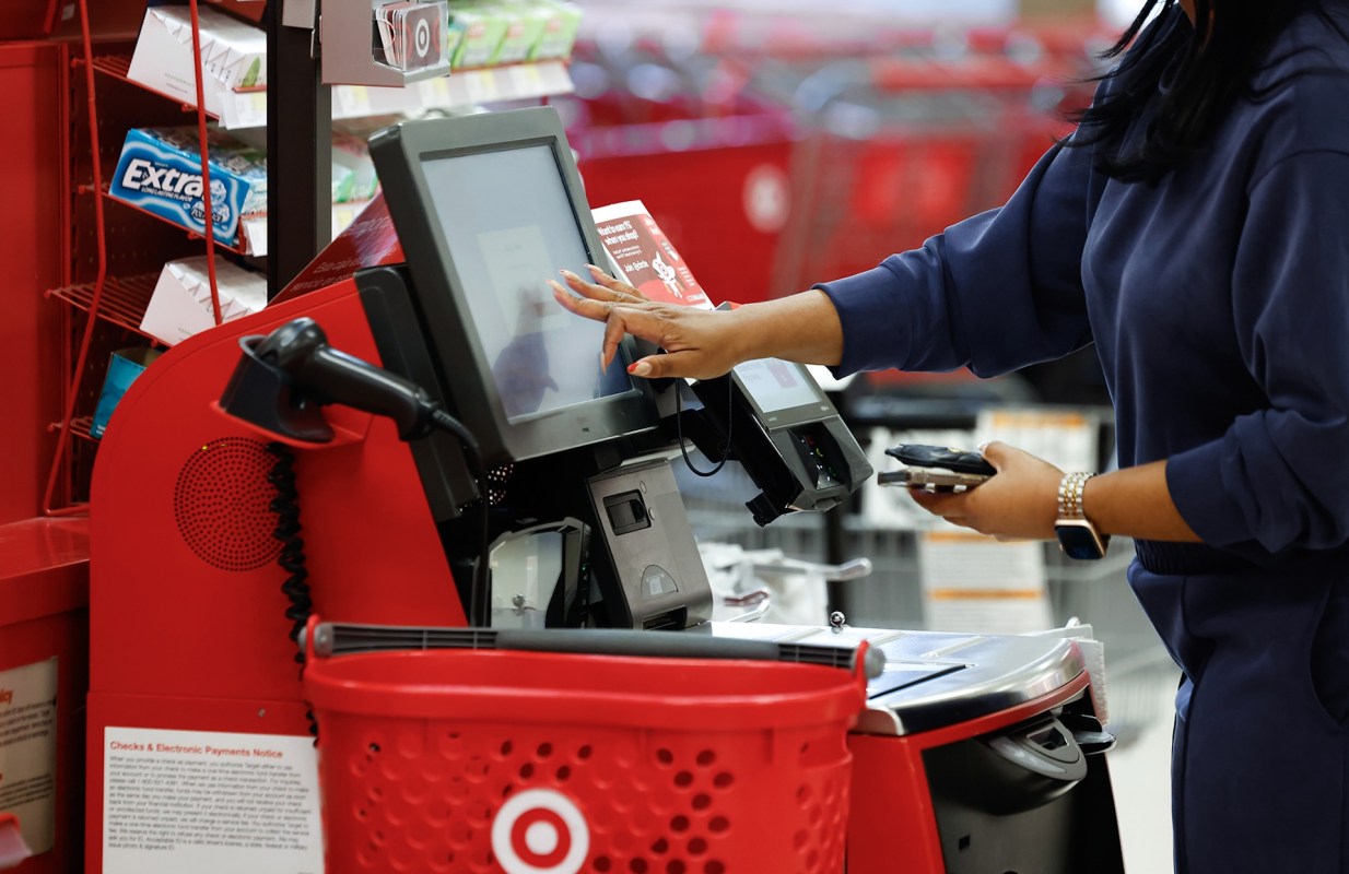A person using a self-checkout kiosk in a Target store, touching the screen with one hand while holding a phone in the other.